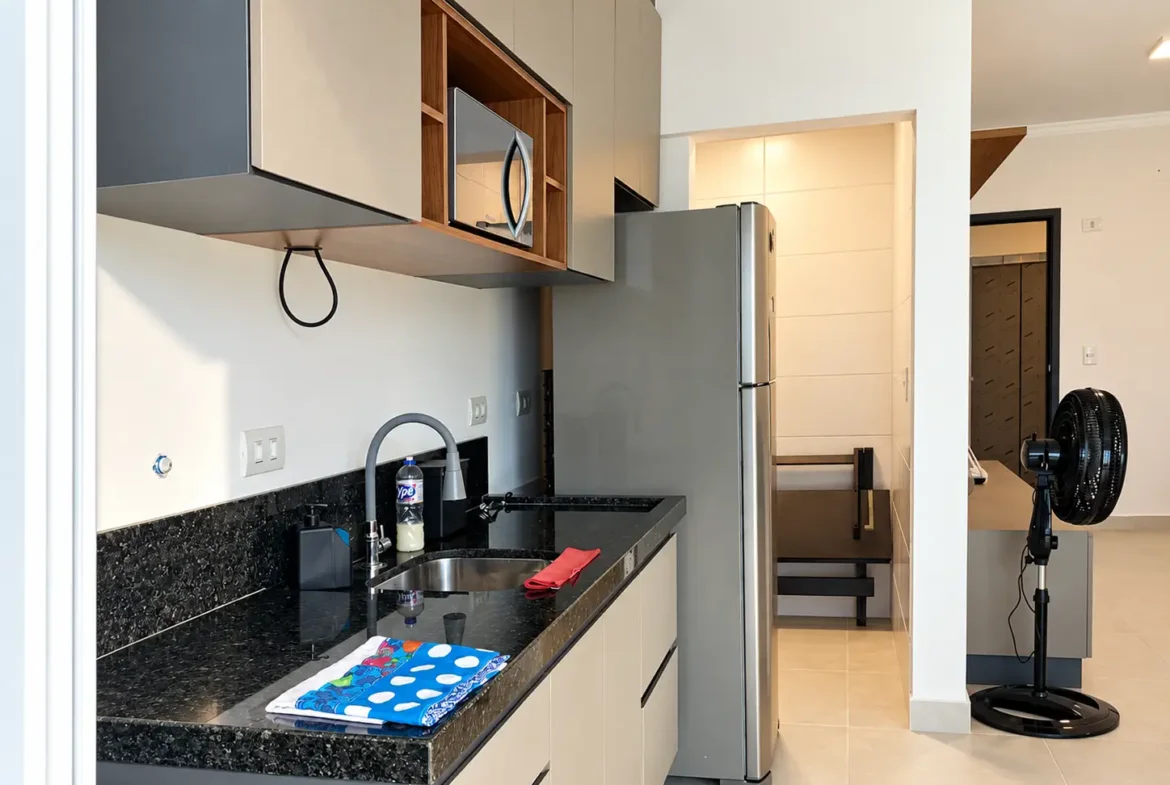 Outro ângulo da cozinha com bancada escura, pia, cooktop e geladeira.