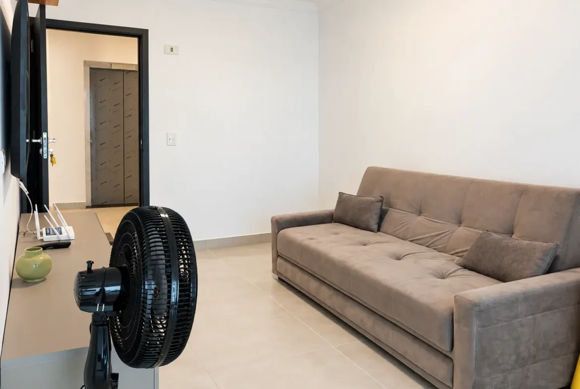 Vista da cozinha integrada à sala de estar, com sofá e ventilador de coluna.