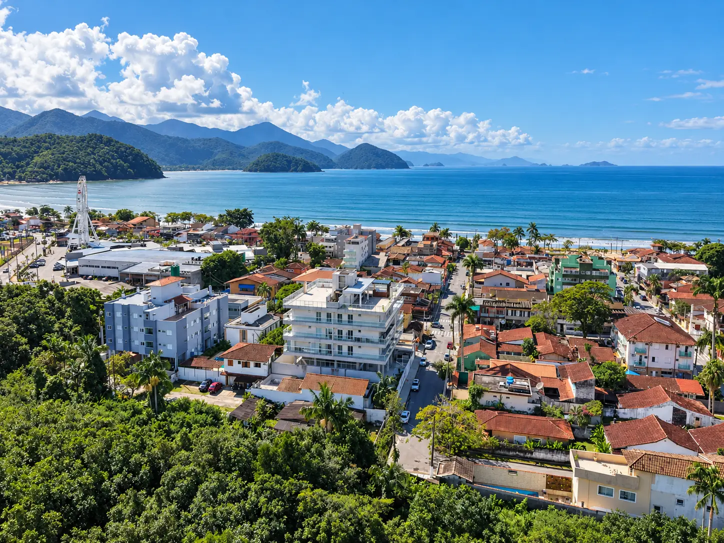 apartamento-itagua-lg117-113-82m2-aerea Vista aérea de região urbana litorânea, com edifícios, vegetação, mar e montanhas ao fundo.