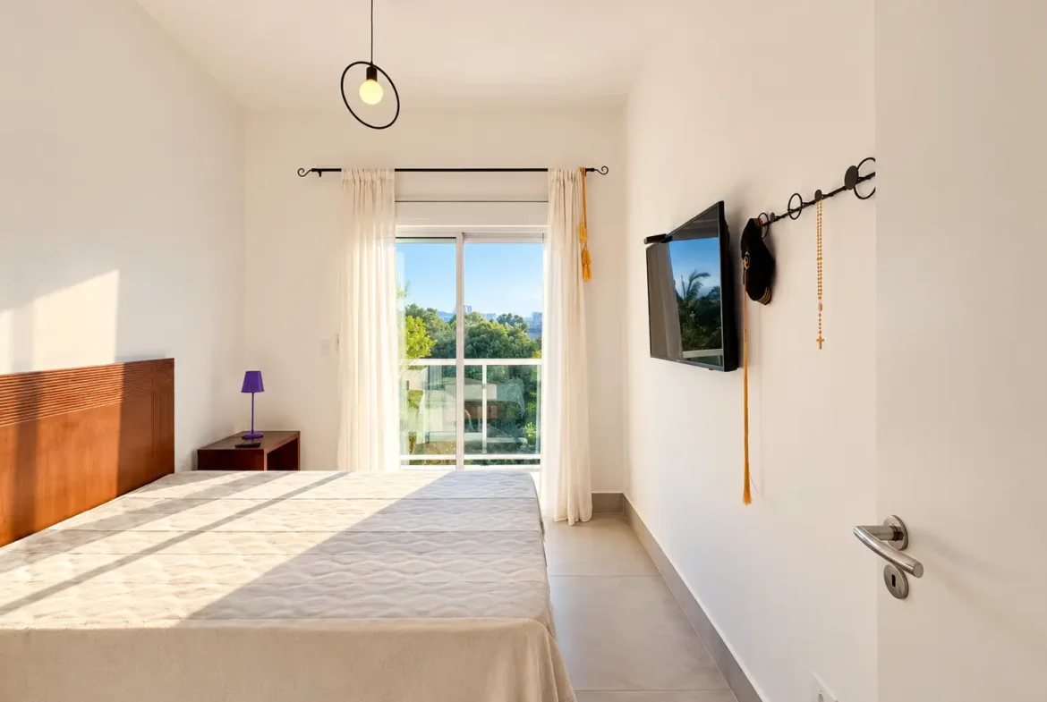 Dormitório com cama, móveis claros, televisão na parede e janela.
