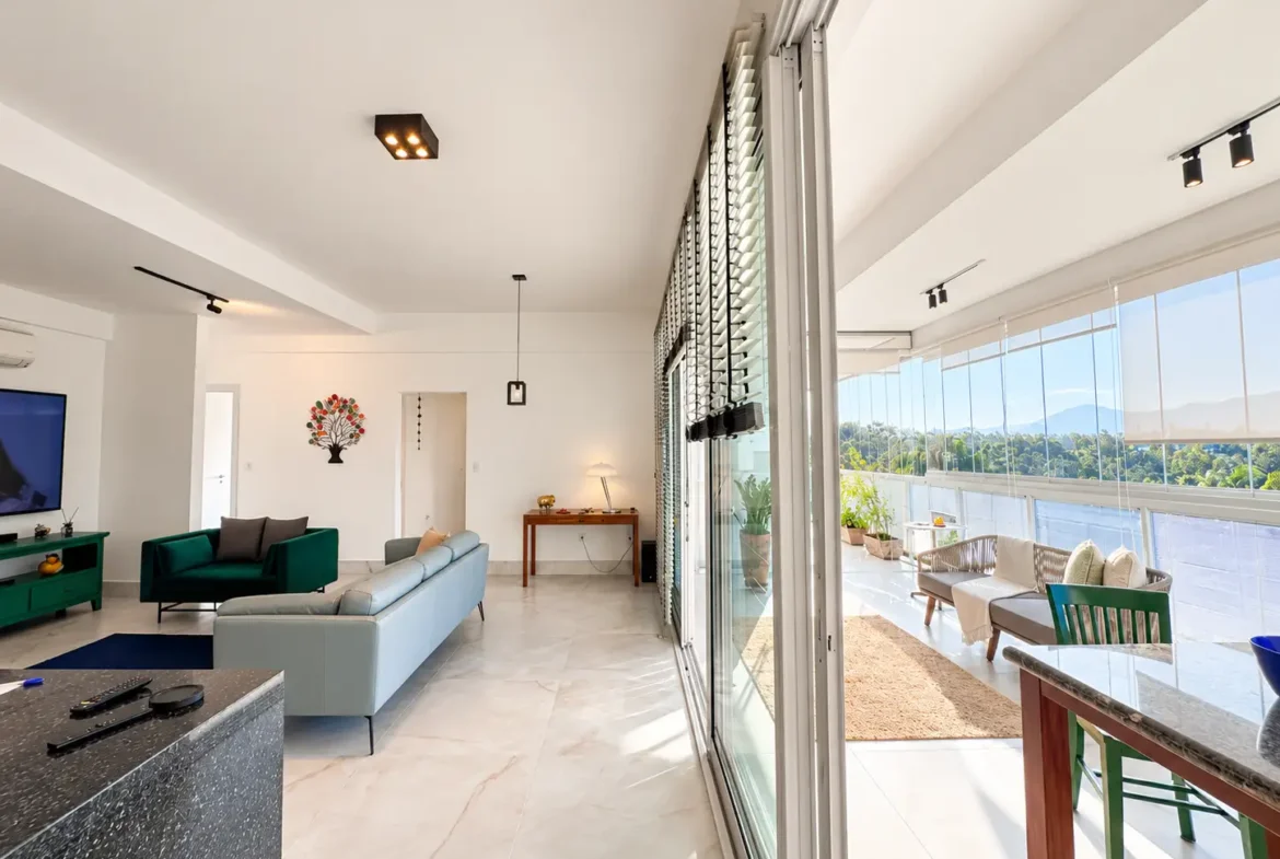 Sala de estar com sofá e televisão, integrada à varanda por grandes portas de vidro.