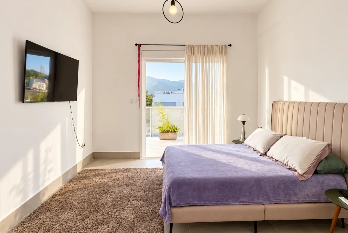 Suíte com cama de casal, roupa de cama lilás e porta de acesso à varanda.