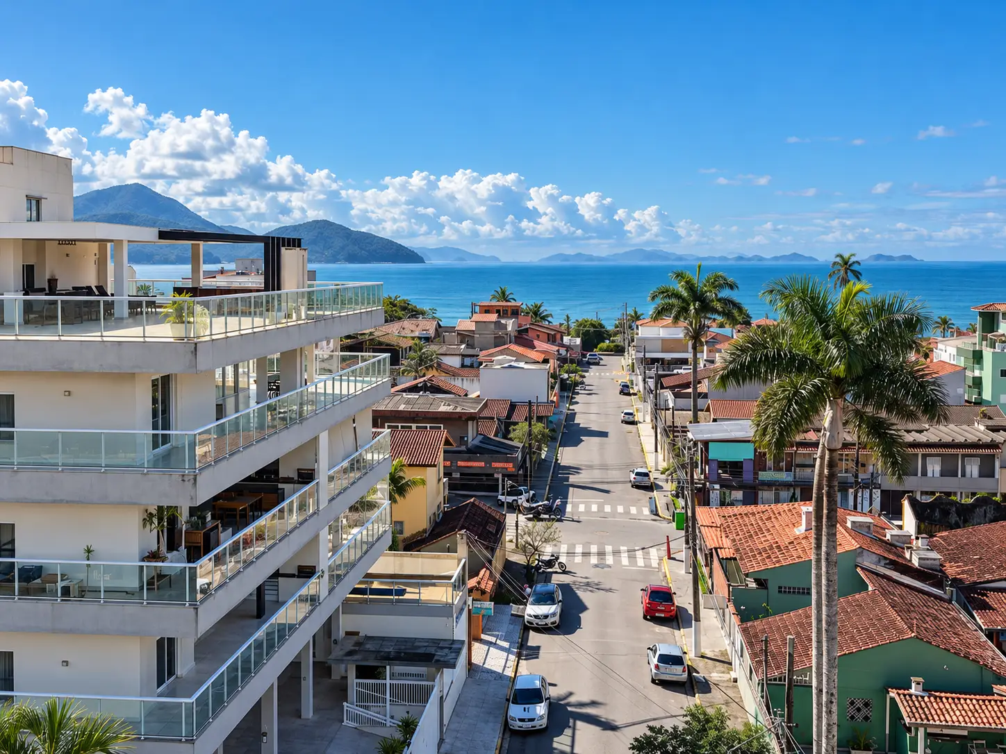 apartamento-itagua-lg117-113-82m2-vista-mar Vista elevada de rua com edifícios e palmeiras, com o mar e montanhas ao fundo.