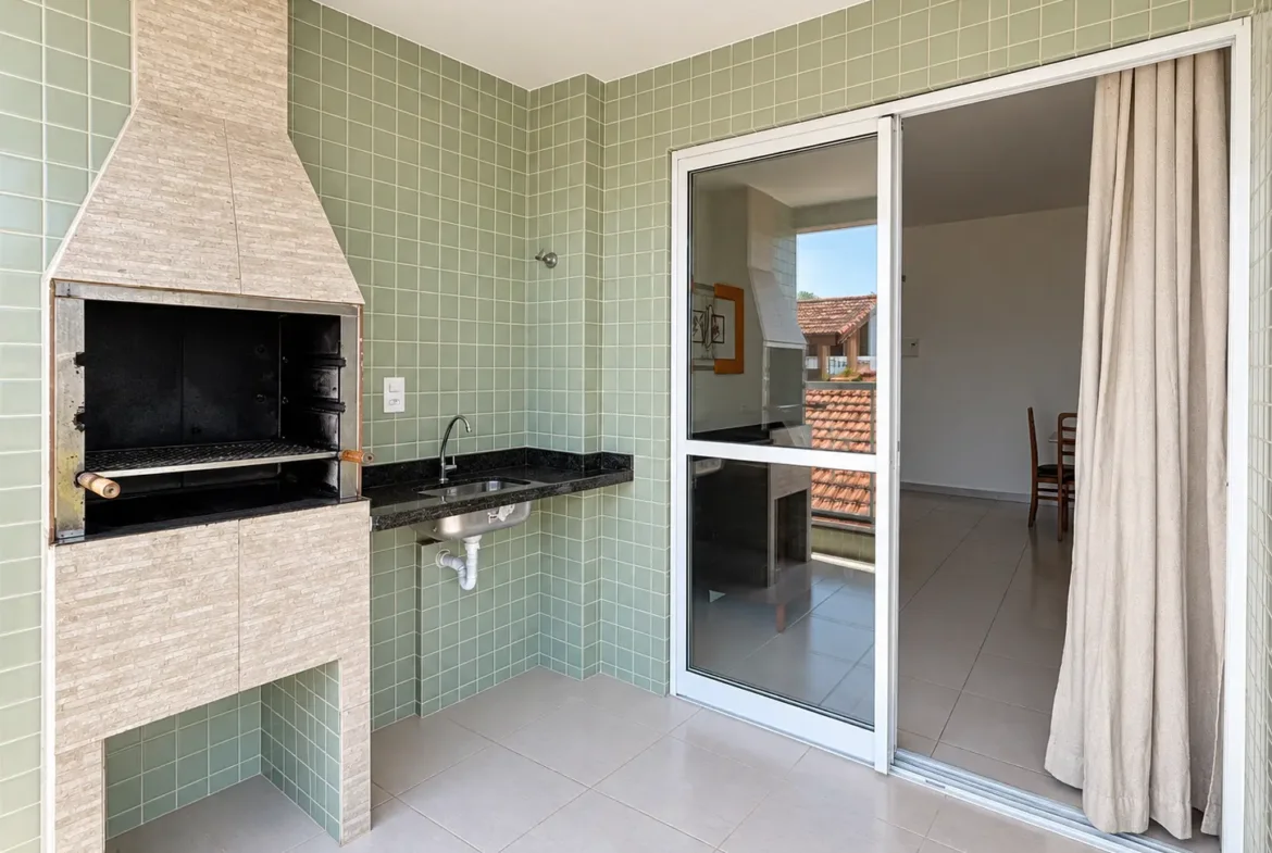 Outro ângulo da varanda gourmet com churrasqueira, bancada e porta de vidro de acesso à sala.
