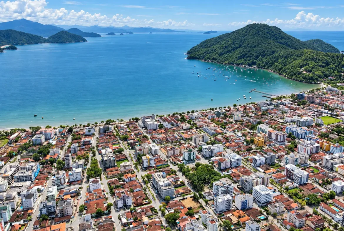 Panorama aéreo da cidade junto ao litoral, com edifícios, vegetação e baía ao fundo.