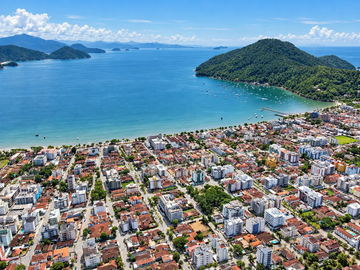 loft-35-m2-Itaguá-varanda-piscina-bairro022 Panorama aéreo da cidade junto ao litoral, com edifícios, vegetação e baía ao fundo.