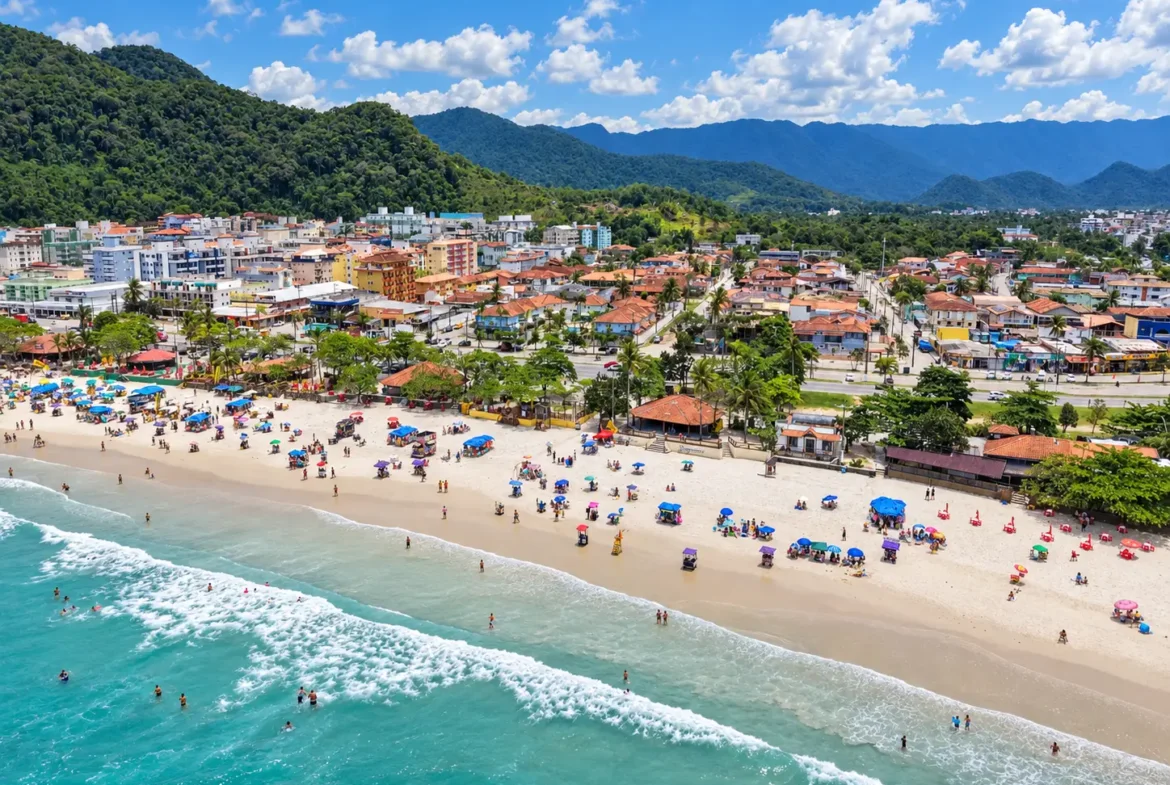 Vista aérea da orla com faixa de areia, ondas, edificações e vegetação ao redor.